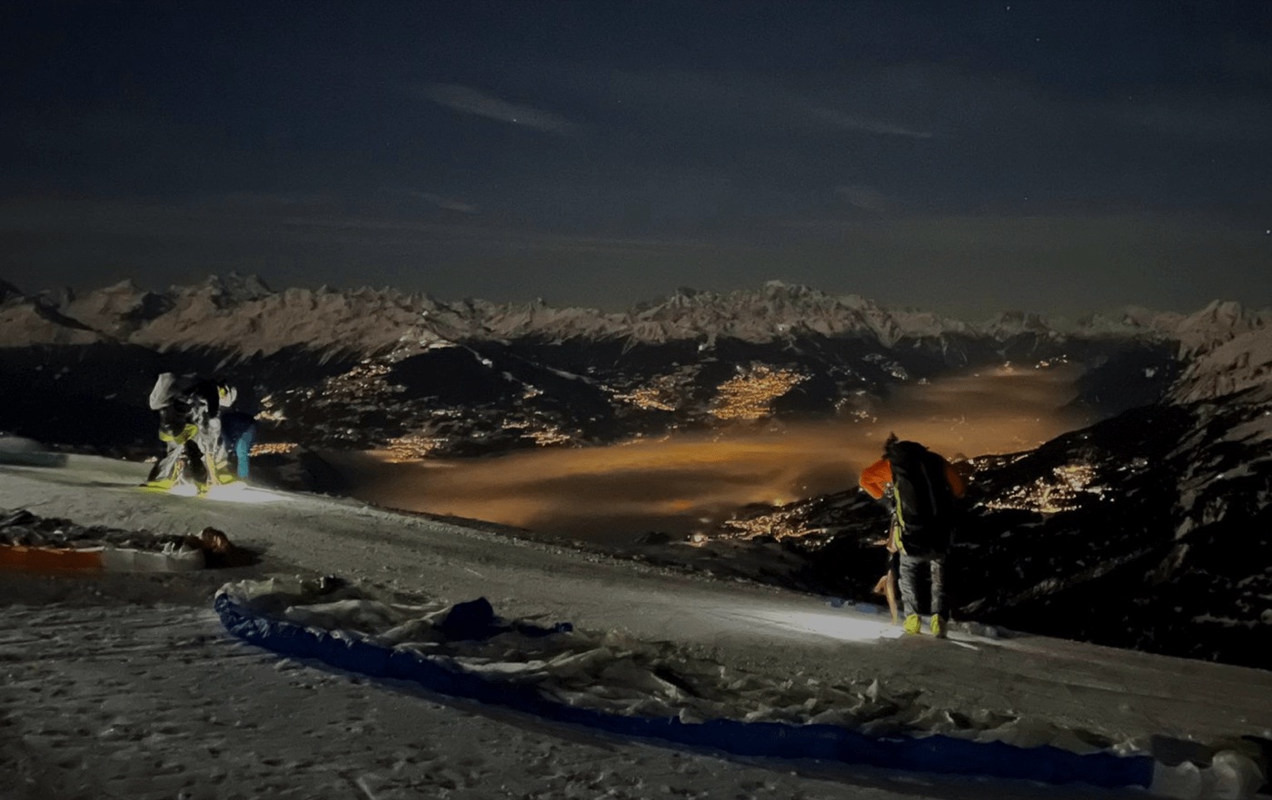 Préparation des parapentes sur la neige de nuit, au-dessus de la vallée du Rhône illuminée