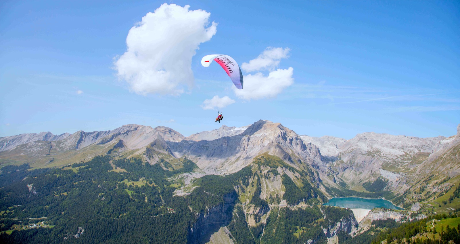 Parapente biplace sous un nuage, au-dessus des sommets et d'un lac de montagne